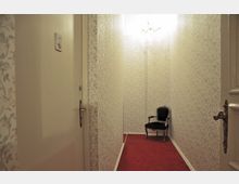 Schmaler Flur mit floraler Tapete, rotem Teppichboden und einer dunklen, gepolsterten Armlehnenstuhls an der Wand; im Vordergrund eine geöffnete Tür.