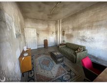 Schlichter, alt wirkender Innenraum mit hohen Decken, rau verputzten und teils fleckigen Wänden sowie einer weißen Tür an der Stirnseite. Im Raum stehen ein gemusterter Teppich, ein grünes Sofa, eine Truhe als Tisch und ein kleines Sideboard mit Lampen, beleuchtet von einer einfachen Deckenleuchte.