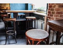 Ein gemütlich eingerichtetes Café mit dunkelbraunen Holzbarhockern und einem Tisch, umgeben von einer gemauerten Wand und einer blauen Wand. Ein Fenster mit Blumen im Hintergrund lässt natürliches Licht herein.