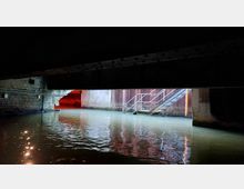 Blick unter eine niedrige Brücke in einen Kanal mit Wasser, beleuchtet durch eine Mischung aus weißem und rotem Licht. Auf der rechten Seite führt eine Metalltreppe ins Wasser, während links eine gemauerte Wand und ein weiterer Zugang sichtbar sind.
