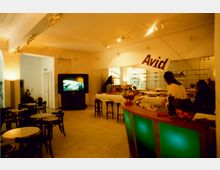 Ein Innenraum mit einem modernen Thekenbereich, beleuchtet durch grüne Lichtelemente, mehreren runden Tischen und Stühlen im Vordergrund und einer großen Leinwand im Hintergrund. Rechts sind Barhocker und ein Buffetbereich sichtbar, darüber hängt ein Banner mit der Aufschrift „Avid“.