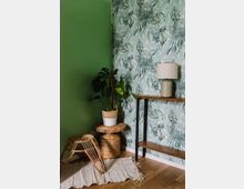 Ecke eines Raumes mit einer tropisch gemusterten Tapete und einer grünen Wand, Einrichtung mit einem Rattanhocker, einem Beistelltisch aus Korbgeflecht, einer Topfpflanze und einer schlichten Lampe auf einem Holztisch mit Metallbeinen, auf einem hellen Teppich.