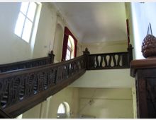 Ein Innenraum mit einer großen Holztreppe, die zu einer Galerie mit verzierten Geländern führt. Die Wände sind hell gestrichen, mit sichtbaren Rissen, und es gibt hohe Fenster sowie rote Vorhänge, die Licht einlassen.