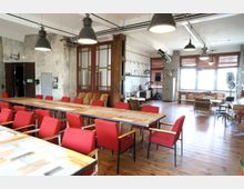 Industrie-inspirierter Veranstaltungsraum mit Holzboden, langen Holztischen und roten Polsterstühlen. Im Hintergrund sind große Fenster, rustikale Möbel, versetzte Beleuchtung und dekorative Elemente sichtbar.