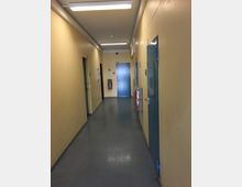 Langer, schmaler Innenflur mit beige gestrichenen Wänden und grauem Boden, beleuchtet durch rechteckige Deckenleuchten. Entlang des Gangs befinden sich mehrere blaue Türen, am Ende eine Tür mit der Nummer „107“ sowie ein Feuerlöscher und ein Wandhydrant.