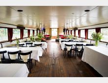 Innenraum eines festlich dekorierten Schiffes mit Holzboden, weißen Tischdecken, schwarzen Stühlen und Blumenschmuck. Große Fenster mit roten Vorhängen lassen Tageslicht herein, und am Ende des Raumes befinden sich grüne Sofas und eine kleine Bühne.