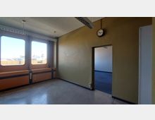 Ein leerer Raum mit gelben Wänden und großen Fenstern, durch die man eine Außenszene mit Gebäuden sehen kann. An der Wand hängt eine analoge Uhr, und durch eine offene Tür ist ein weiterer Raum mit grauem Teppichboden sichtbar.