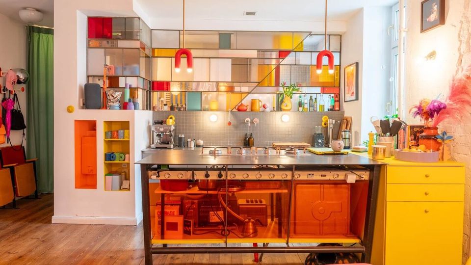 Bright, modern kitchen with a stainless-steel island and orange-tinted glass base, lit by two red pendant lights. A wall of open shelving with multicolored glass panels forms a backdrop above a gray tile backsplash, with a yellow chest of drawers and wooden floor in the foreground.