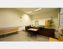 Ein kleiner, funktionaler Gerichtssaal mit schlichtem Mobiliar, darunter ein hölzernes Richterpult, ein Tisch mit Stuhl und eine Holzbank. Der Raum hat helle Wände, eine Tür mit abgerundetem Rahmen und ein großes Fenster, durch das Tageslicht einfällt.