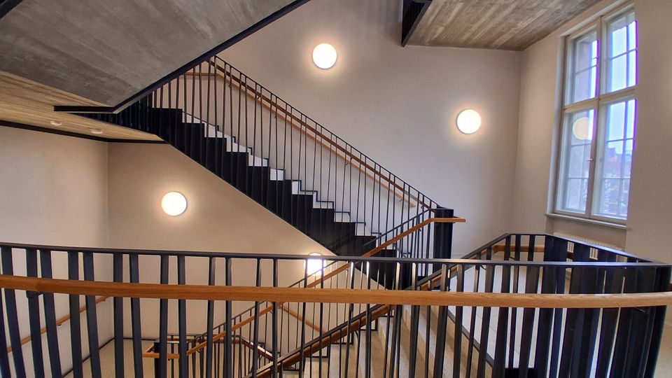 Innenansicht eines modernen Treppenhauses mit mehrläufiger Treppe, schwarzen Metallgeländern und Holzhandläufen. Helle Wände mit runden Wandlampen und ein großes Fenster an der rechten Seite lassen Tageslicht in den Raum.