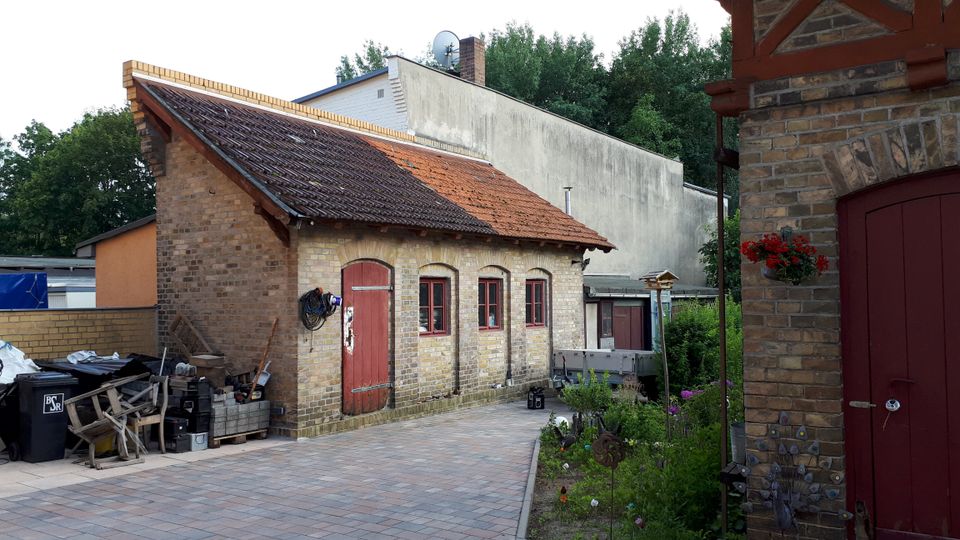 Innenhof mit gepflasterter Fläche vor einem kleinen Backsteingebäude mit Satteldach aus roten Ziegeln, roten Holztüren und mehreren kleinen Fenstern. Rechts ist der Rand eines weiteren Backsteinbaus mit Fachwerkdetails und roter Tür zu sehen, daneben ein schmaler Gartenstreifen mit Pflanzen.