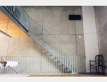 Innenraum mit hohen Sichtbetonwänden und einer geraden Metalltreppe mit Geländer, die diagonal zu einer oberen Ebene führt. Links ist eine Holzdecke zu sehen; rechts hängen ein Bildschirm und ein Ausgangsschild, daneben steht ein einzelner Holzstuhl.