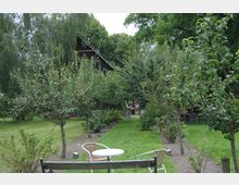 Garten mit Obstbäumen und Rasenflächen, im Vordergrund stehen Tische und Stühle aus Holz und Metall, im Hintergrund ist ein dunkles Holzhaus von Bäumen umgeben.