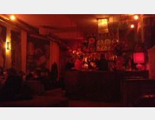 Eine gemütliche Bar mit warmem, rötlichem Licht, einem zentralen Tresen mit Blumenarrangements und dekorativen Elementen. Im Hintergrund sind gemusterte Tapeten sowie Sitzbereiche mit gedämpfter Beleuchtung sichtbar.