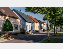 Reihenhäuser mit Satteldächern in einer ruhigen Wohnstraße, gesäumt von Bäumen und Gehwegen; die Häuser verfügen über Vorgärten mit Zäunen und vereinzelten Bäumen.