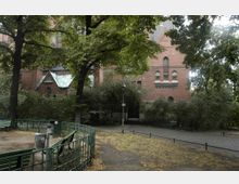 Backstein-Gebäude mit gotischen Fenstern und einem begrünten Metallschindeldach, umgeben von Bäumen und Sträuchern. Im Vordergrund befinden sich ein kleiner gepflasterter Platz mit einer Laterne, Grünflächen mit Zaun sowie Sitzbänke.