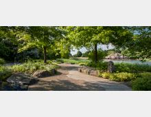 Ein Parkweg mit gepflastertem Boden führt durch eine grüne Landschaft, gesäumt von niedrigen Pflanzen, Büschen und großen Steinen. Im Hintergrund sind ein See sowie eine strohgedeckte Hütte mit Sonnenschirmen sichtbar, umgeben von Bäumen.