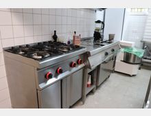 Industrieküche mit Edelstahlgeräten, darunter ein Gasherd mit vier Kochfeldern und roten Drehknöpfen, sowie eine Arbeitsfläche mit eingebautem Mixer und weiteren Küchenutensilien. Die Wände sind mit weißen Fliesen verkleidet, und der Raum hat ein gefliestes Grau-Bodenmuster.