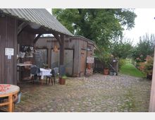 Hofgelände mit rustikalen Holzhäusern und einem gepflasterten Innenhof. Vor einem Gebäude steht ein kleiner Tisch mit weißer Tischdecke und zwei Stühlen, umgeben von Blumentöpfen und Grünflächen.