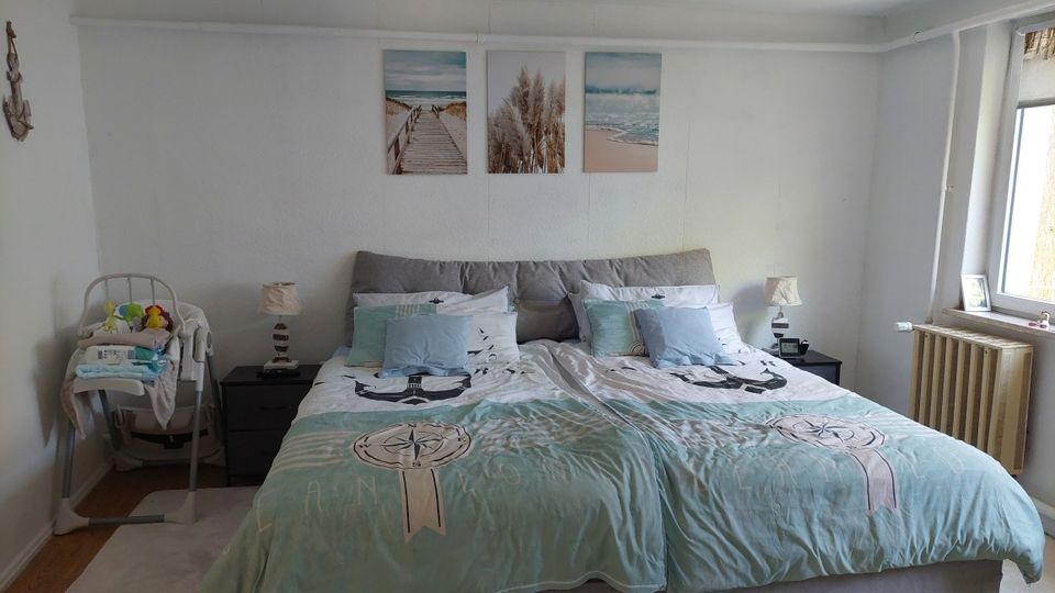 Schlafzimmer mit großem Doppelbett und hellblauer Bettwäsche, flankiert von zwei Nachttischen mit Lampen; darüber hängen drei gerahmte Strandmotive. Rechts befindet sich ein Fenster mit Heizkörper darunter, links steht ein Baby-Hochstuhl mit abgelegten Textilien.