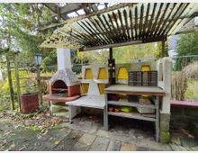 Außenbereich mit einem überdachten Grillplatz aus Beton, bestehend aus einem gemauerten Grill, einer Arbeitsfläche mit Regalen darunter und gelben Glaselementen in der Rückwand. Im Hintergrund sind Bäume, Sträucher und ein Maschendrahtzaun zu sehen.