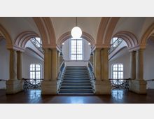 Großzügiger Treppenaufgang in einem historischen Gebäude mit hohen Marmorsäulen, verzierten schmiedeeisernen Geländern und Rundbögen. Im Hintergrund befindet sich ein großes Fenster, das natürliches Licht in den Raum lässt.