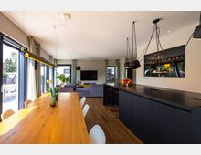 Moderne Wohnküche mit offenem Grundriss, einem großen Holzesstisch links, einer schwarzen Kücheninsel rechts und einer lila Sofaecke im Hintergrund. Große Fenster lassen viel Tageslicht herein, und elegante Hängeleuchten ergänzen die Einrichtung.