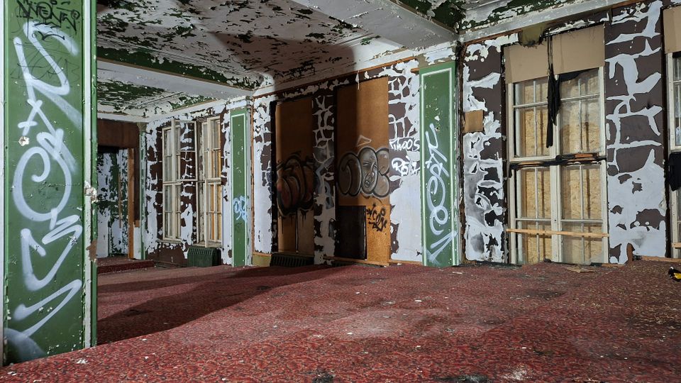 Innenraum eines verlassenen Gebäudes mit abblätternder Farbe an Decke und Wänden, grün-weiß gestrichenen Säulen und mehreren hohen Fenstern, teils mit Brettern vernagelt. Der Raum hat roten Teppichboden und ist mit Graffiti und beschädigten Wandverkleidungen übersät.