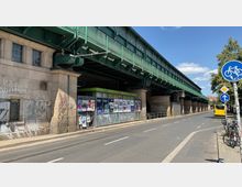 Straßenansicht unter einer stählernen Eisenbahnbrücke mit grüner Bemalung, daneben ein kleines Gebäude mit Graffiti und Plakaten an den Wänden. Rechts daneben verläuft eine Straße mit Fahrbahnmarkierungen, Fahrrädern und einem Bus im Hintergrund.