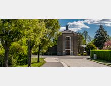 Kleine Kirche mit schlichtem rechteckigen Baukörper, Satteldach und einem Kreuz auf dem Dachfirst; im Vordergrund eine asphaltierte Straße, flankiert von Bäumen und gepflegten Hecken. Der Eingangsbereich der Kirche ist durch ein hohes, rechteckiges Fenster mit Holzstreben hervorgehoben.