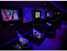 Eine moderne Lounge mit schwarzen Ledersofas und niedrigen Tischen, beleuchtet von violettem und blauem Licht. An den Wänden hängen Bildschirme mit leuchtenden digitalen Kunstmotiven, und auf den Tischen stehen Sektkübel und Gläser.