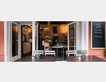 Außenbereich eines kleinen Cafés oder Restaurants mit Holztischen und Stühlen, dekoriert mit kleinen Topfpflanzen. Durch offene Glastüren ist der Innenraum mit einer Bar und gedeckten Tischen sichtbar, daneben eine Tafel mit handgeschriebenem Menü.