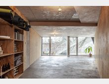 Minimalistisch gestalteter Innenraum mit Sichtbetondecke, Holzwänden und großflächigen Fenstern, die auf eine Außentreppe und einen begrünteren Hof blicken. Links befinden sich offene Holzregale mit aufgerollten Matten, Stoffen und Utensilien, rechts steht eine Topfpflanze vor den Fenstern.