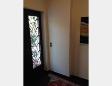 Eingangsbereich mit heller Wand und dunklen Fliesenboden. Links eine Tür mit einem dekorativen Glaselement und Metallgitter, rechts ein farbiges Wandbild. Ein gemusterter Teppich liegt auf dem Boden.