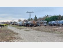Ein Abstellgleis mit mehreren Güterwaggons, hauptsächlich Kesselwagen, die in Reihen geparkt sind. Im Hintergrund befinden sich Bäume, ein Strommast und ein bewölkter Himmel; der Boden besteht aus Kopfsteinpflaster und unbefestigtem Gras.