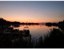 Sonnenuntergang über einem stillen See mit vereinzelten Booten am Ufer und Schilf im Vordergrund. Der Himmel ist in warmen Orange- und Blautönen gefärbt, während Bäume und Silhouetten im Hintergrund die Szenerie einrahmen.