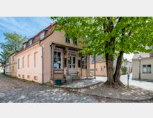 Ein kleines, pastellfarbenes Gebäude mit roten Ziegeldachgauben und einem hölzernen Eingangsbereich, beschriftet mit "Wein Duval", steht an einer gepflasterten Straße. Vor dem Eingang stehen ein Baum, ein Fass und eine Straßenlaterne, während rechts und links angrenzende Häuser sichtbar sind.