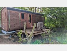 Holzhütte auf einem Anhänger mit schwarzem Schindeldach, Fenster und Tür, davor eine erhöhte Holzterrasse mit Stufen und einfachen Holzmöbeln. Die Hütte steht in einer grünen, von Bäumen und Sträuchern umgebenen Garten- oder Waldrandumgebung.
