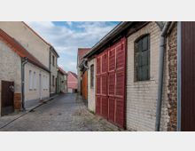 Schmale, gepflasterte Gasse mit niedrigen Wohn- und Nebengebäuden aus unterschiedlichen Baumaterialien, darunter verputztes Mauerwerk und Ziegel. Rechts im Vordergrund eine weiß gemauerte Wand mit roten Holztoren und einem schmalen Fenster; links hell gestrichene Häuser mit roten Ziegeldächern.