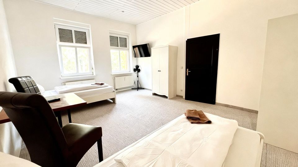 Helles, schlichtes Gästezimmer mit zwei Einzelbetten, grauem Teppichboden und weißer Holzdecke. Zwei Fenster mit Rollläden, ein kleiner Tisch mit Stuhl, ein Schrank sowie ein wandmontierter Fernseher und eine dunkle Tür sind zu sehen.