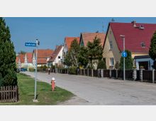 Straßenszene in einem Wohngebiet mit Reihen von Einfamilienhäusern mit roten und orangefarbenen Satteldächern auf beiden Seiten. Ein Straßenschild mit der Aufschrift „Gaußstraße“ ist vorne sichtbar, begleitet von Bäumen, Vorgärten und Zäunen entlang der Gehwege.