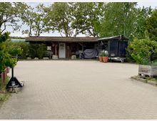 Holzgebäude mit überdachter Veranda, umgeben von Bäumen und Pflanzen. Der Bereich davor besteht aus einem gepflasterten Hof mit vereinzelten Blumenkübeln und Holzkisten.