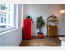 Ecke eines Wohnraums mit einem roten Retro-Kühlschrank, einer großen Zimmerpflanze in einem Topf und einem schlichten Regal aus Rattan. Links befindet sich ein großes Fenster, das Tageslicht hereinlässt, und rechts hängt eine minimalistische Deckenleuchte.