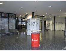 Eingangsbereich eines modernen Gebäudes mit glänzendem dunklem Boden, Sitzbänken, Glaswänden und Infotafeln. Im Hintergrund befinden sich ein Treppenaufgang, ein Aufzug und eine Wand mit Türen sowie Zeitschriftenregale.