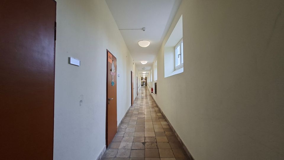 Langer, schmaler Innenflur mit hellem Anstrich, gefliestem Boden und einer Reihe von Türen auf der linken Seite. Rechts befinden sich kleine Fenster und Wandnischen, die Decke ist mit runden Leuchten entlang des Gangs ausgestattet.