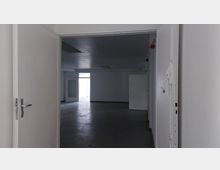 Ansicht eines leeren, rechteckigen Raumes mit weiß gestrichenen Wänden und grauem Boden, durch den leicht geöffneten Türrahmen fotografiert. Im Hintergrund sind geschlossene Türen, ein Fenster mit Glasfläche und Deckenleuchten sichtbar.