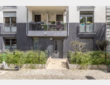 Mehrfamilienhaus mit moderner Fassade aus dunklen Klinkersteinen und weißen Putzflächen, Balkonen mit Blumenkästen und einem zentralen Eingang mit Hausnummer „95“. Vor dem Gebäude befinden sich niedrige Hecken, ein Gehweg aus Pflastersteinen und ein abgestellter E-Scooter.