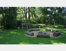 Waldlichtung mit einer Feuerstelle, umgeben von Baumstämmen als Sitzgelegenheiten. Im Hintergrund befindet sich ein gemauerter Grill vor einer dichten Baum- und Strauchkulisse.