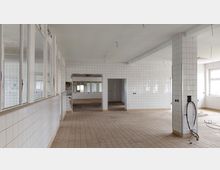 Leerer Innenraum eines ehemaligen Gebäudes mit gefliesten Wänden und Böden, mehreren Fenstern und offenen Durchgängen. Die Decke zeigt lose Kabel, und es sind keine Möbel oder Geräte vorhanden.