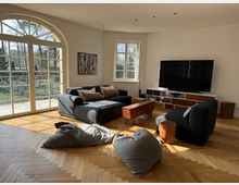 Wohnzimmer mit großen, hellen Fenstern, die einen Blick auf einen Garten bieten. Der Raum enthält ein graues Sofa, einen Sessel, Sitzkissen, einen Fernseher auf einem bunten Sideboard und hat einen Boden aus Holzparkett in Fischgrätmuster.